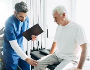 Younger male therapist holding a clipboard. He is examining the right knee of an older man with white hair who is sitting on an exam table.