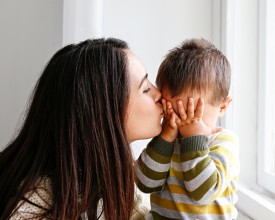 Young mother with long brown hair kissing the cheek of a small boy who is wearing a striped shirt and covering his eyes with his hands