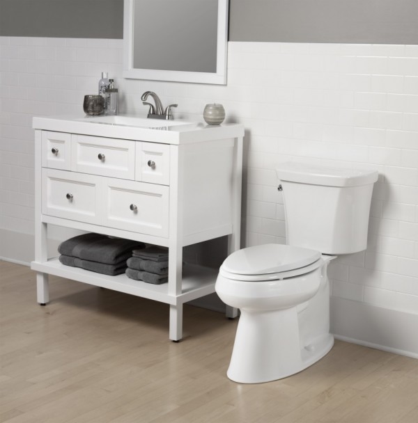 White toilet next to small white vanity with grey towels on bottom shelf and mirror on wall