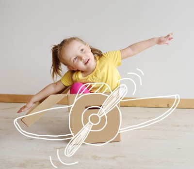 Toddler girl wearing yellow shirt sitting in cardboard box pretending it's an airplane