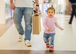 Father wearing jeans carrying paper bag. Toddler girl wearing striped red shirt and jeans is walking next to him.