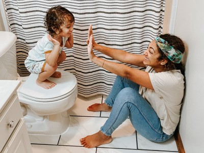 Mother sitting on bathroom floor high fiving toddler son crouched on toilet bowl lid