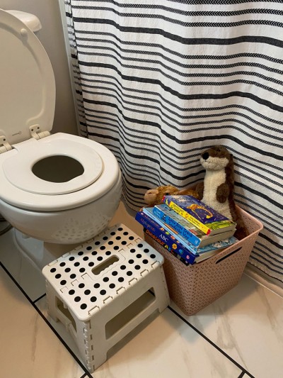 Toilet with potty seat installed next to box of books and black striped shower curtain