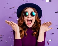 Young woman wearing hat and sunglasses, mouth open in surprise with hands up