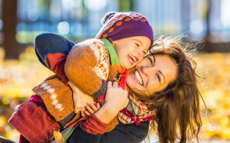 Smiling mother wearing sweater hugging a small child outside in fall setting with leaves