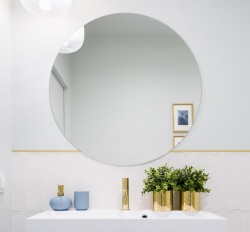 Large round mirror over white vanity with blue soap dispenser and potted plants in gold containers