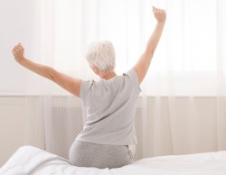 Back view of older woman wearing pajamas sitting on bed with arms outstretched