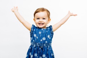 Toddler girl with red hair and a big smile, wearing blue dress and holding her arms up in happiness