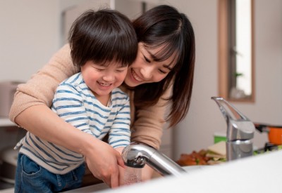 Smiling mother with arms around toddler helping her wash hands at sink
